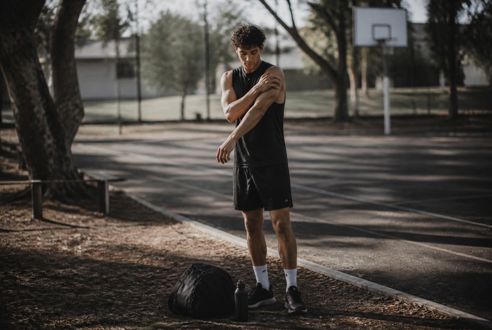 Recreational athlete in dark training gear packing up beside a suburban Australian outdoor court, calmly rolling a shoulder after a session in soft morning light